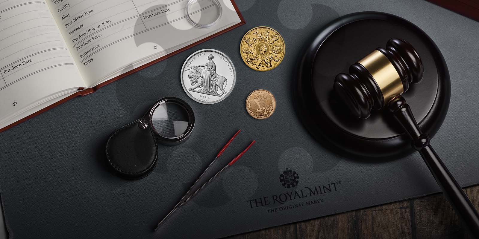 Three coins, a dark wood gavel and striking block, foldable magnifying glass, mahogany coin storage box, tweezers, and log book sitting on a coin mat embossed with The Royal Mint The Original Maker.