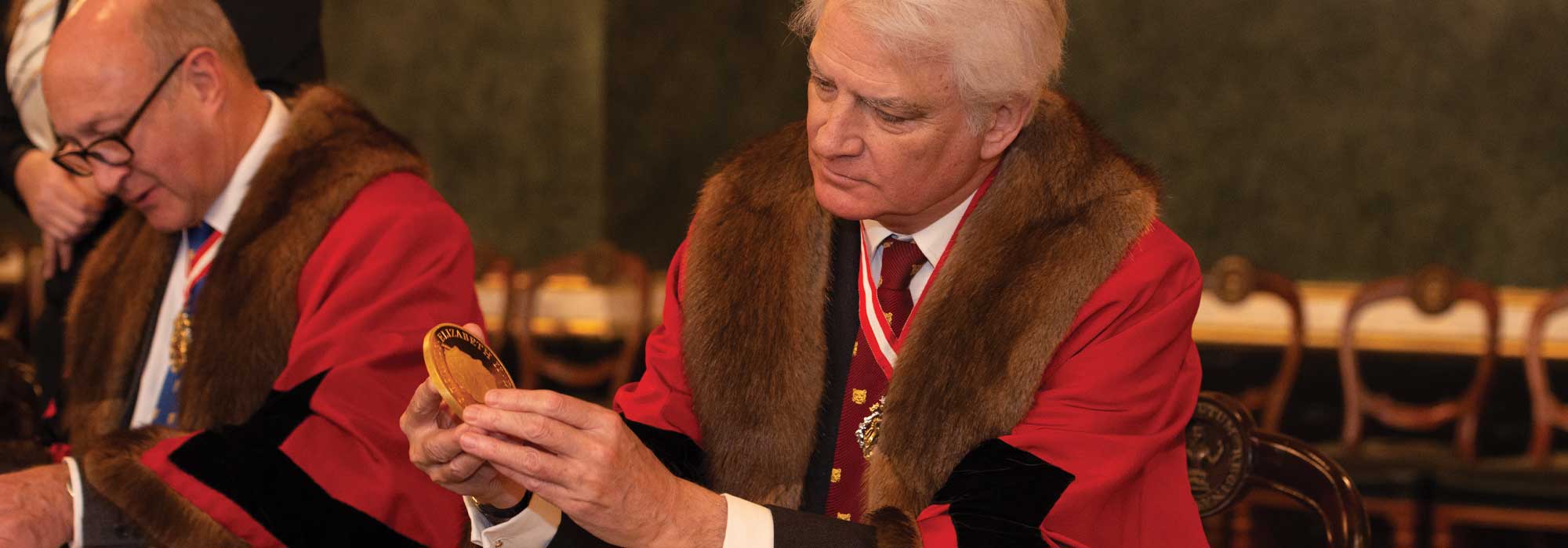 A photograph of a member of the jury in traditional dress inspecting a large gold coin at the historic Trial of the Pyx ceremony.