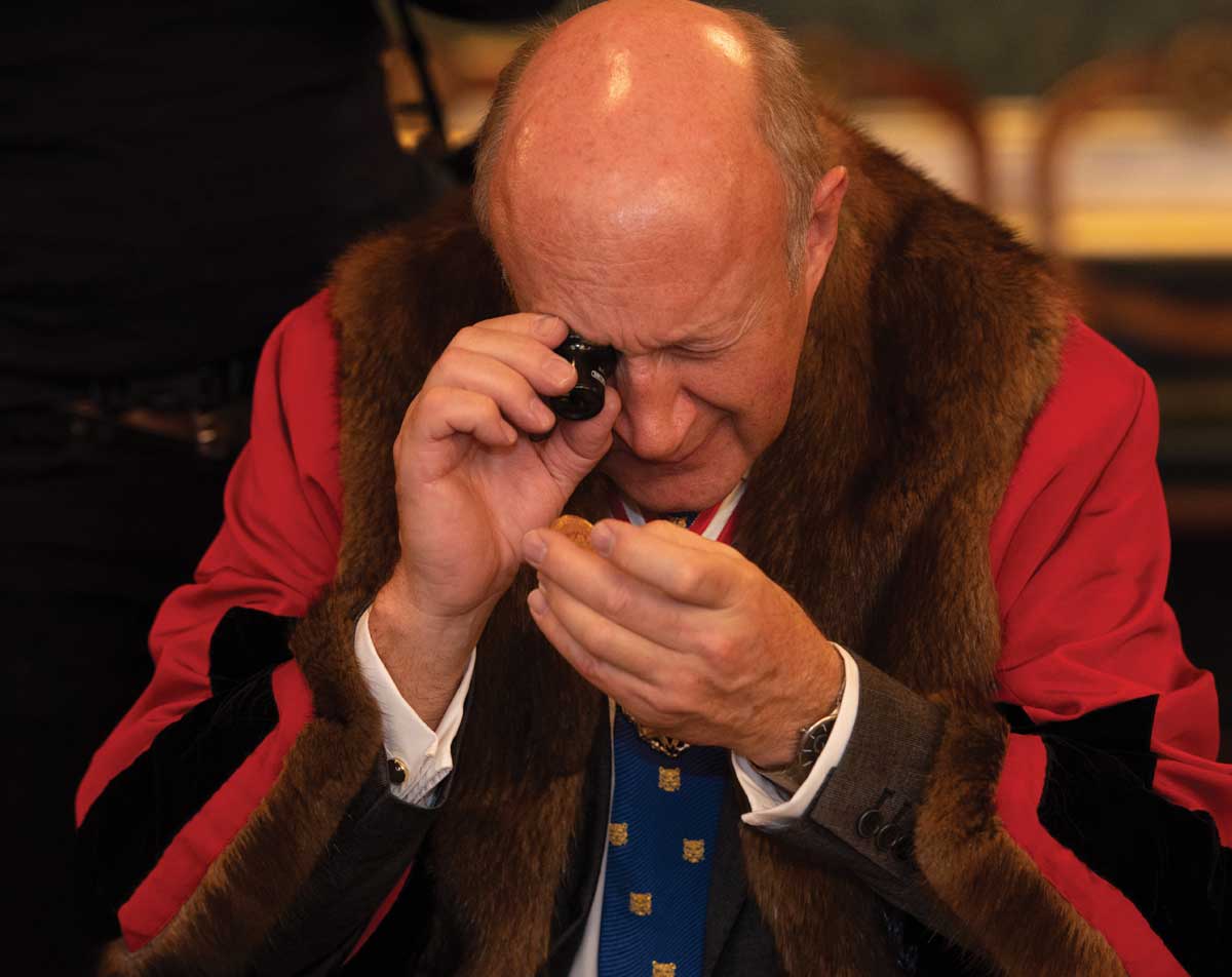 A photograph of a man in traditional dress inspecting a gold coin in his hand at the Trial of the Pyx ceremony.