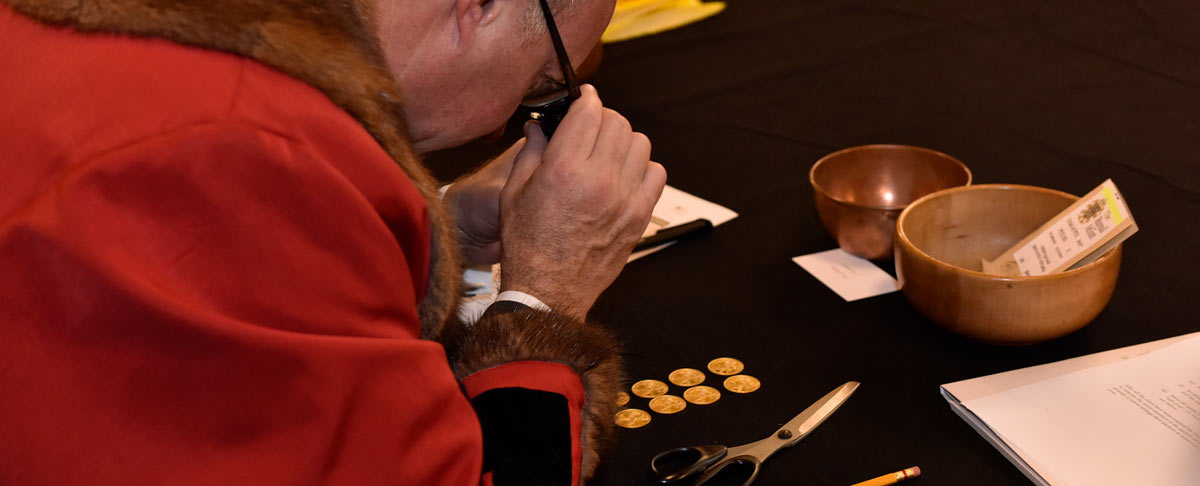 A photograph of a man in traditional dress inspects gold coins with a microscope at the historic Trial of the Pyx ceremony.